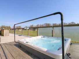 An outdoor hot tub on a wooden deck with glass railing and a grassy field in the background at Angel Lodge - No 27 in Fornham St Genevieve near Bury St Edmunds
