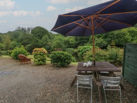 An outdoor seating area by a pond with a wooden table, two metal chairs, an umbrella, and greenery at Lazy Trout Caravan in Ludchurch near Narberth