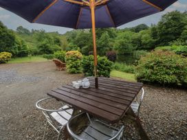 An outdoor wooden table with metal chairs and a large umbrella with tea set near a pond and green trees at Lazy Trout Caravan Ludchurch near Narberth