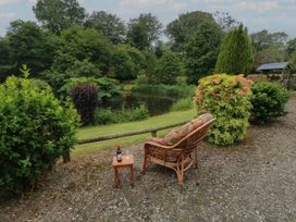 An outdoor seating area with a wicker chair and small wooden table by a pond surrounded by trees and bushes at Lazy Trout Caravan Ludchurch near Narberth