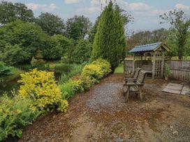 A garden area with wooden benches and a wooden swing near a pond surrounded by green trees and bushes at Lazy Trout Caravan in Ludchurch near Narberth