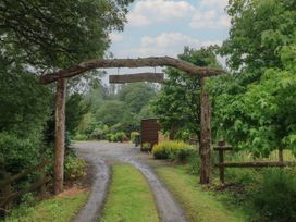 A gravel driveway with grass strips under a wooden archway with a hanging sign surrounded by trees at Lazy Trout Caravan in Ludchurch near Narberth