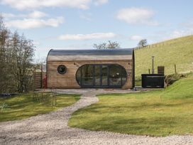 A wooden cabin with round windows and glass doors on a grassy area with a gravel path and a hot tub beside it at Binsey Lodge Longlands near Caldbeck