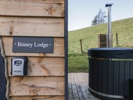 A wooden wall with a Binsey Lodge sign a code lock box and part of an outdoor hot tub with a chimney on a deck near a grassy hill at Binsey Lodge in Longlands near Caldbeck