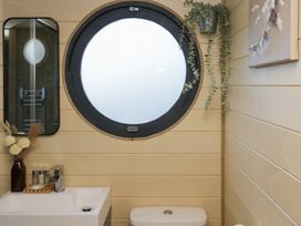 A bathroom with a round window above a toilet a rectangular mirror a sink with a faucet and toiletries and a hanging plant at Binsey Lodge Longlands near Caldbeck