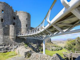 A stone castle with a modern white metal bridge leading to its entrance surrounded by grass and rocky terrain at Pen Y Bryn Bach in Harlech