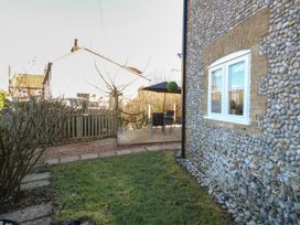 A garden with grass and bushes next to a stone house with a wooden patio and outdoor furniture at Cobble Cottage in Great Ryburgh