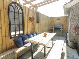An outdoor patio area with wooden benches and table along a wooden fence with cushions and a wall mirror and a barbecue grill and potted plants at Ty Copr in Amlwch
