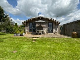 A wooden cabin with two glass doors a patio with a table umbrella and chairs surrounded by grass and plants at Artisan Lodge in Longridge