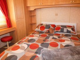 A bedroom with a bed featuring patterned bedding a wooden wardrobe a small red stool and a window with a red curtain at Meric Stone Holiday Lets Caravan 2 Benone near Castlerock