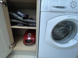 A washing machine next to an open cabinet with a red vacuum cleaner and hose inside at 12 Thurlestone Rock in Thurlestone