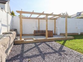 A backyard with a wooden pergola and wooden seating area next to a white wall and gravel and grass at Trearddur House Cottage in Trearddur Bay