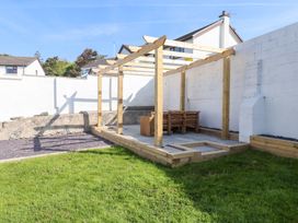 A garden area with grass a wooden pergola with seating and a white wall at Trearddur House Cottage in Trearddur Bay