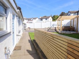 A paved walkway beside a white exterior wall with windows and a door next to a wooden fence and a wooden pergola with benches in a backyard at Trearddur House Cottage in Trearddur Bay
