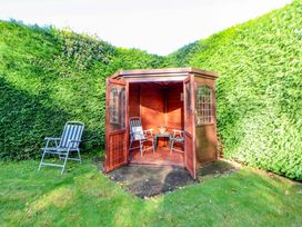 A small wooden garden shed with open doors containing two chairs and a table next to a hedge with a chair on the lawn