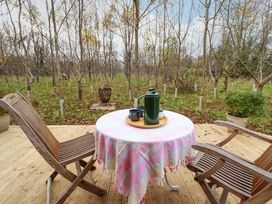 A wooden deck with two wooden chairs and a round table covered with a pink checkered tablecloth with a green thermos and cups on the table in a garden with young trees at Hobhole in Keal Cotes near East Kirkby
