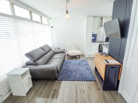 A living room with a gray sofa a white armchair a blue patterned rug a wooden cabinet a wall-mounted TV and kitchen area at 104 The Laurels in Chapel St Leonards