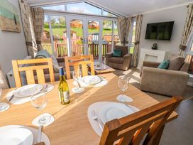 A dining area with a wooden table set with plates glasses and a bottle of wine next to a living room with armchairs a couch a wall-mounted TV and glass doors overlooking a garden at Dragonfly in Tunstall near Hipswell
