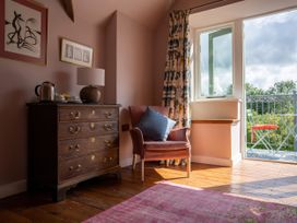 A room with a wooden chest of drawers with a lamp and kettle on top an armchair with a cushion floral curtains and an open door to a balcony with a chair at Coach House Llanwrtyd Wells
