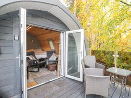 A small cabin with open doors showing a bed and chairs inside and outdoor chairs with a table on a deck surrounded by trees at Elleric Pods - The Last Resort Fasnacloich near Appin