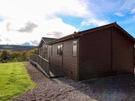 A brown wooden cabin with multiple windows on a gravel and grass lawn with trees and hills in the background at Crathie Lodge in Banavie near Caol
