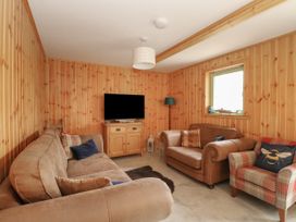 A living room with wooden panel walls a television on a wooden cabinet a brown sectional sofa two armchairs and a floor lamp at Bradan Lodge in Dalmally