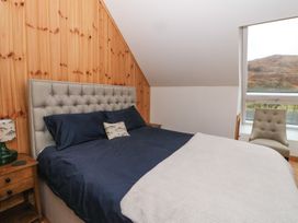 A bedroom with a bed with blue bedding a wooden bedside table with a lamp a beige chair and a window showing hills at Bradan Lodge in Dalmally