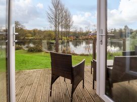 View through open glass doors onto a wooden deck with wicker chairs overlooking a lake and grassy area at Lakeview Lodge in Forton near Garstang