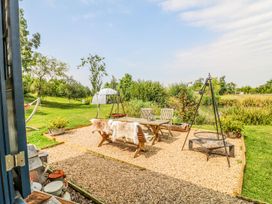 An outdoor seating area with wooden table and benches with fur covers on a gravel surface near greenery and a hammock at Lillies Shepherds Hut at Shepherds Lodge Bottesford near Redmile and Vale of Belvoir