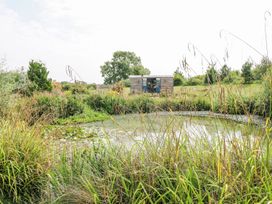 A pond surrounded by tall grass with a small wooden hut and trees in the background at Lillies Shepherds Hut at Shepherds Lodge Bottesford near Redmile and Vale of Belvoir