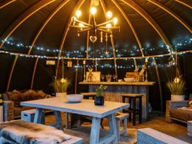 An interior of a wooden hut with a central table, benches, a bar counter with stools, decorative lights, and rustic furniture at Lillies Shepherds Hut at Shepherds Lodge Bottesford near Redmile and Vale of Belvoir