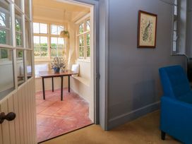 A small sunroom with a wooden table and potted plant next to built-in bench seating with cushions adjacent to a room with a blue armchair and a framed peacock painting on the wall at The Gables in Spaxton near Nether Stowey