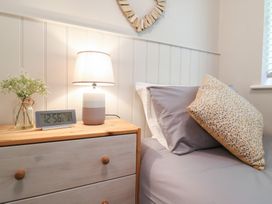 A bedside table with a lamp an alarm clock and flowers next to a bed with pillows at 1 Kessingland Cottages in Kessingland