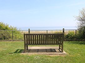 A wooden bench on grass facing a chain link fence with bushes and a beach beyond at 1 Kessingland Cottages in Kessingland