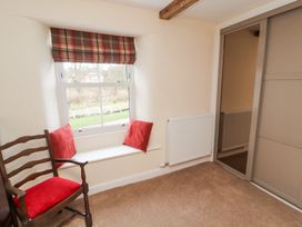 A room with a wooden chair with red cushion next to a window seat with red pillows and a plaid window blind at 1 Brookside in Warcop