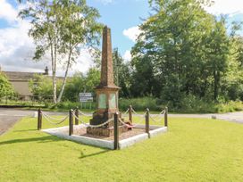 A war memorial with metal chains around it on a grassy area near road signs and trees at 1 Brookside in Warcop