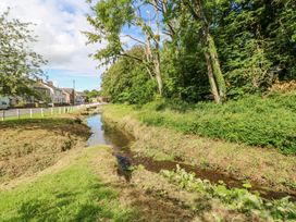 A narrow stream running alongside a row of houses and a road with white fences and trees on the opposite side at 1 Brookside in Warcop