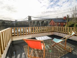 An outdoor terrace with two chairs a table and a patterned rug overlooking a suspension bridge and red brick houses at Ty Chwarel in Menai Bridge