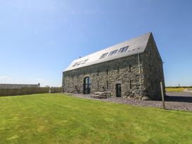 An exterior view of a stone barn with a pitched roof and skylights next to a grassy lawn and a wooden fence at Tithe Barn in Capel Mawr near Malltraeth