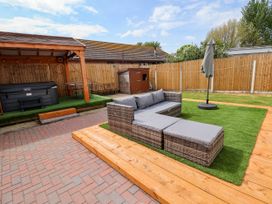 An outdoor patio area with a wicker sofa set on artificial grass and a covered hot tub with chairs behind a wooden fence at Brimrod Talacre