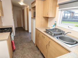 A narrow kitchen with wooden cabinets a stove a small refrigerator a sink with a dish rack and a window showing outdoor buildings at Dunlin Walk 13 in Hayling Island
