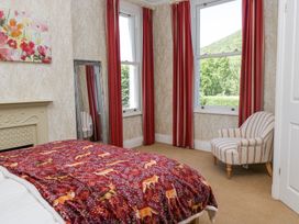 A bedroom with a bed covered in a red animal print blanket a striped chair near two windows with red curtains a tall mirror on the wall and wallpaper with a beige pattern at Sunnycroft in Keswick