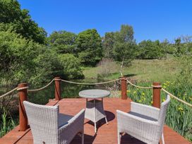An outdoor wooden deck with two wicker chairs and a round table overlooking a pond surrounded by trees and greenery at Celyn in Bontddu near Dolgellau