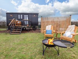 A black shepherds hut on stilts with stairs leading to a deck and outdoor wooden chairs with cushions around a fire pit at Shepherds Hut - Oak in Temple Bruer near Lincoln
