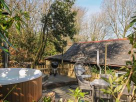An outdoor wooden deck with a round hot tub a patio table with four chairs a hanging chair with cushions and a barbecue grill at Rose Cottage in Callington