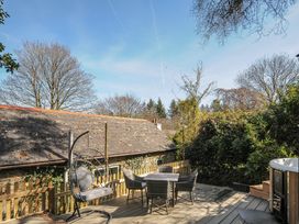 An outdoor deck with a hanging chair, table and chairs set, wooden fence, and hot tub surrounded by trees and a stone building at Rose Cottage in Callington