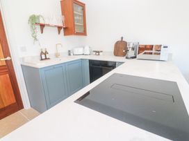 A kitchen with blue lower cabinets a black stovetop a wooden shelf with glasses and plant a wooden cabinet and countertop appliances at Little Victory in Pendeen