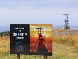 A sign reading welcome to Geevor tin mine with an image of a mining structure at sunset and a view of grass and another mining structure in the background at Little Victory in Pendeen