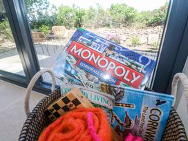A wicker basket with a monopoly Cornwall edition board game magazines and a pink and orange knitted item near a glass door with outdoor chairs and garden visible at Little Victory in Pendeen