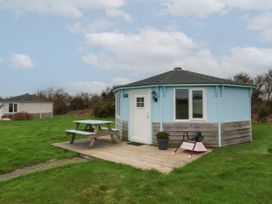 A small round blue and wood cabin with a picnic table and a pink wheelbarrow on a wooden deck surrounded by grass at Shipload Bay near Hartland